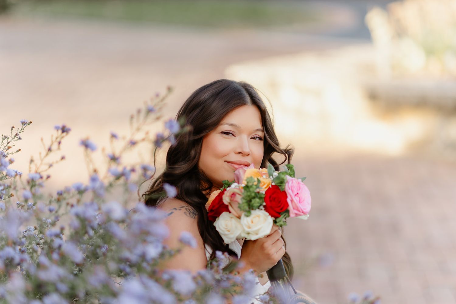 Young woman smiling and holding a colorful bouquet of roses surrounded by purple wildflowers in golden hour light.