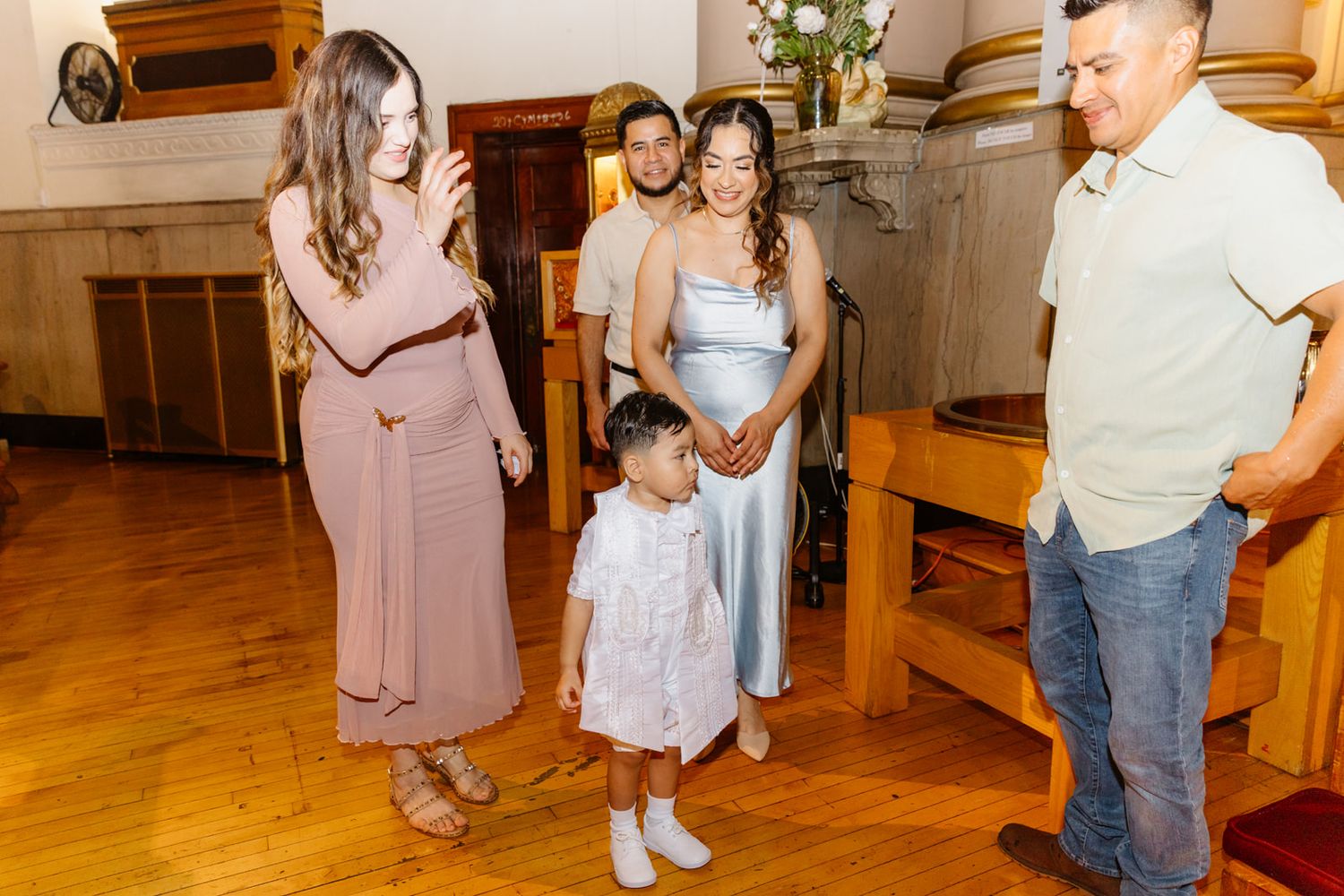 Family with young child in white baptism outfit gathered with priest inside a beautiful church.