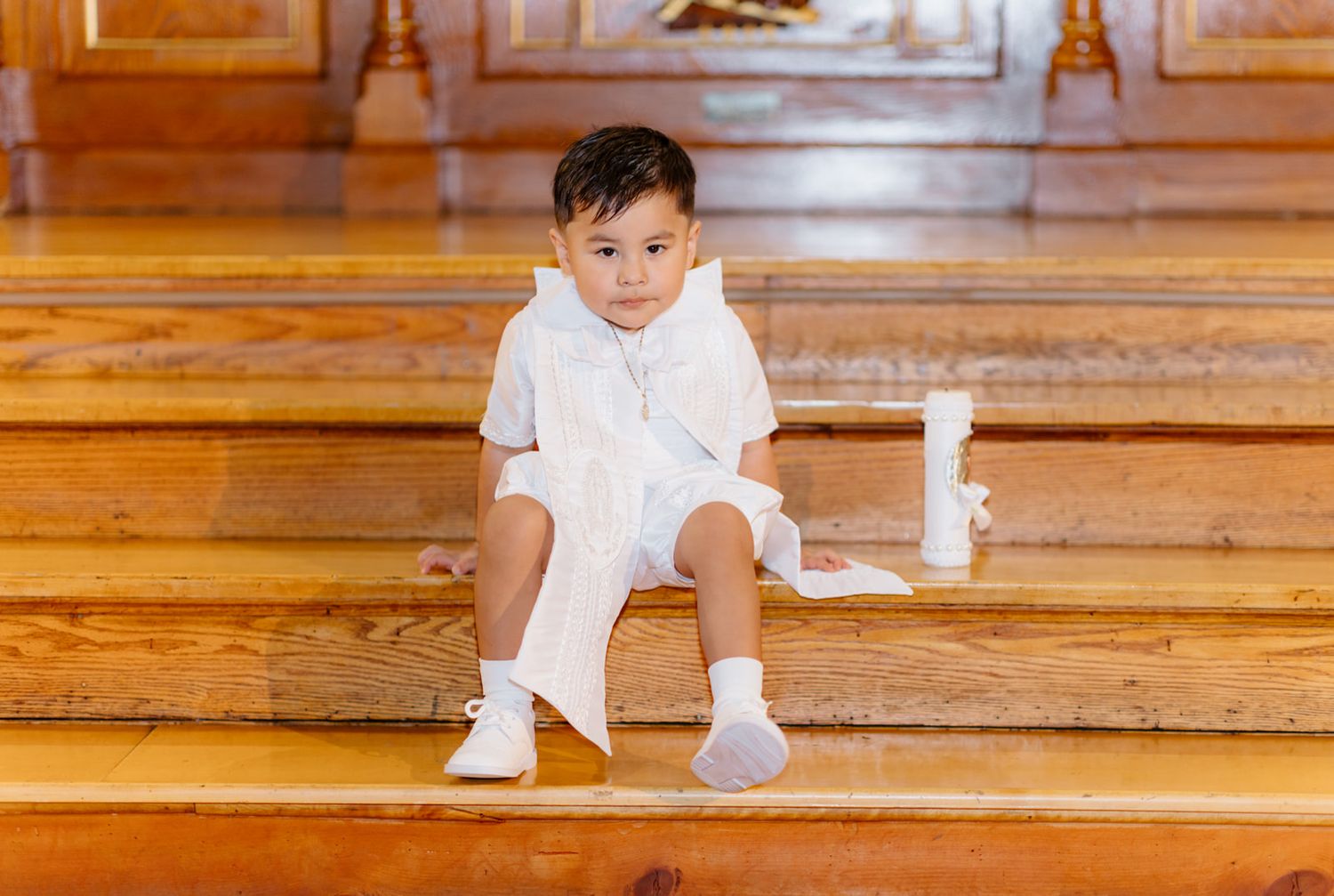 Young boy in white outfit sitting on wooden church pews holding a candle during a baptism ceremony.