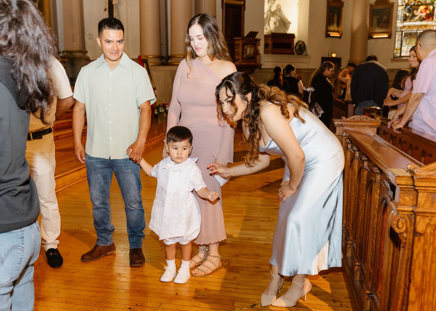 Family with toddler in white dress at Catholic church baptism reception on hardwood floors.
