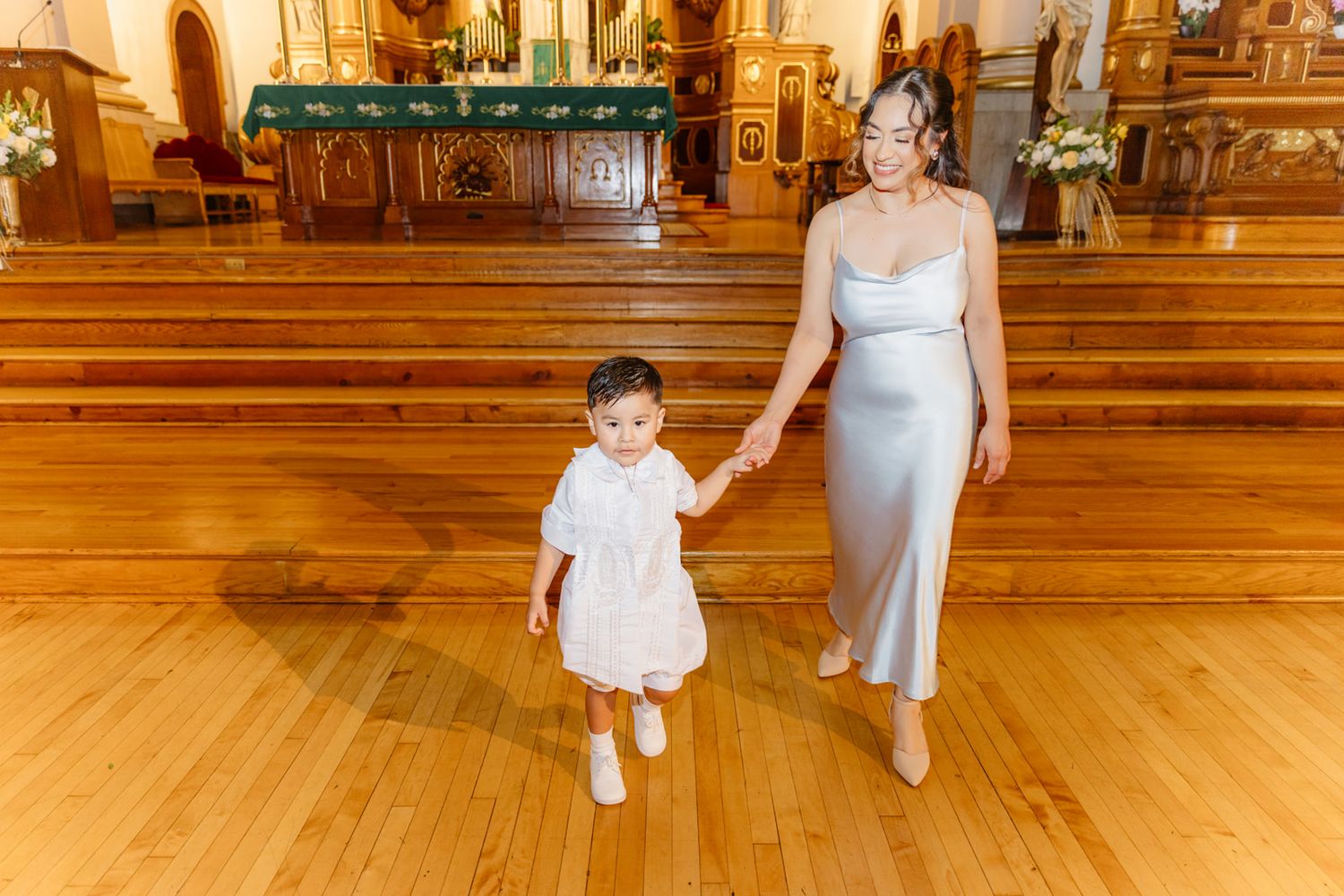 Mother in silver gown walks hand-in-hand with toddler in white dress on church floor.