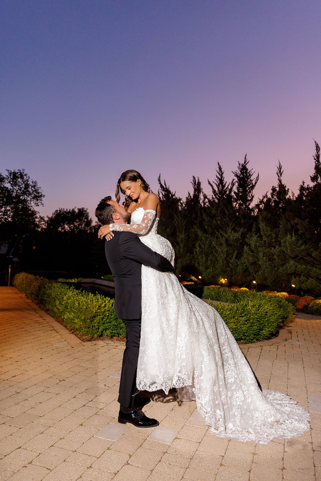 Groom lifts bride in a lace ball gown on a garden pathway during a stunning purple twilight sunset.