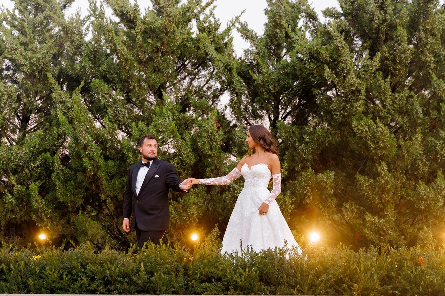 Bride and groom hold hands in lush garden with warm lights, bride in white gown, groom in black tuxedo.