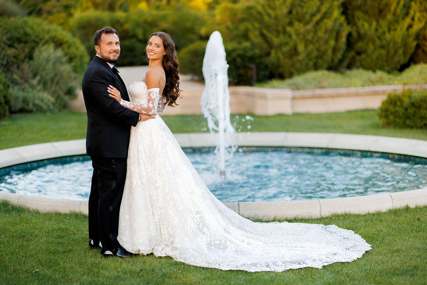 Smiling newlyweds pose together by garden fountain, bride in off-shoulder lace gown with flowing train.