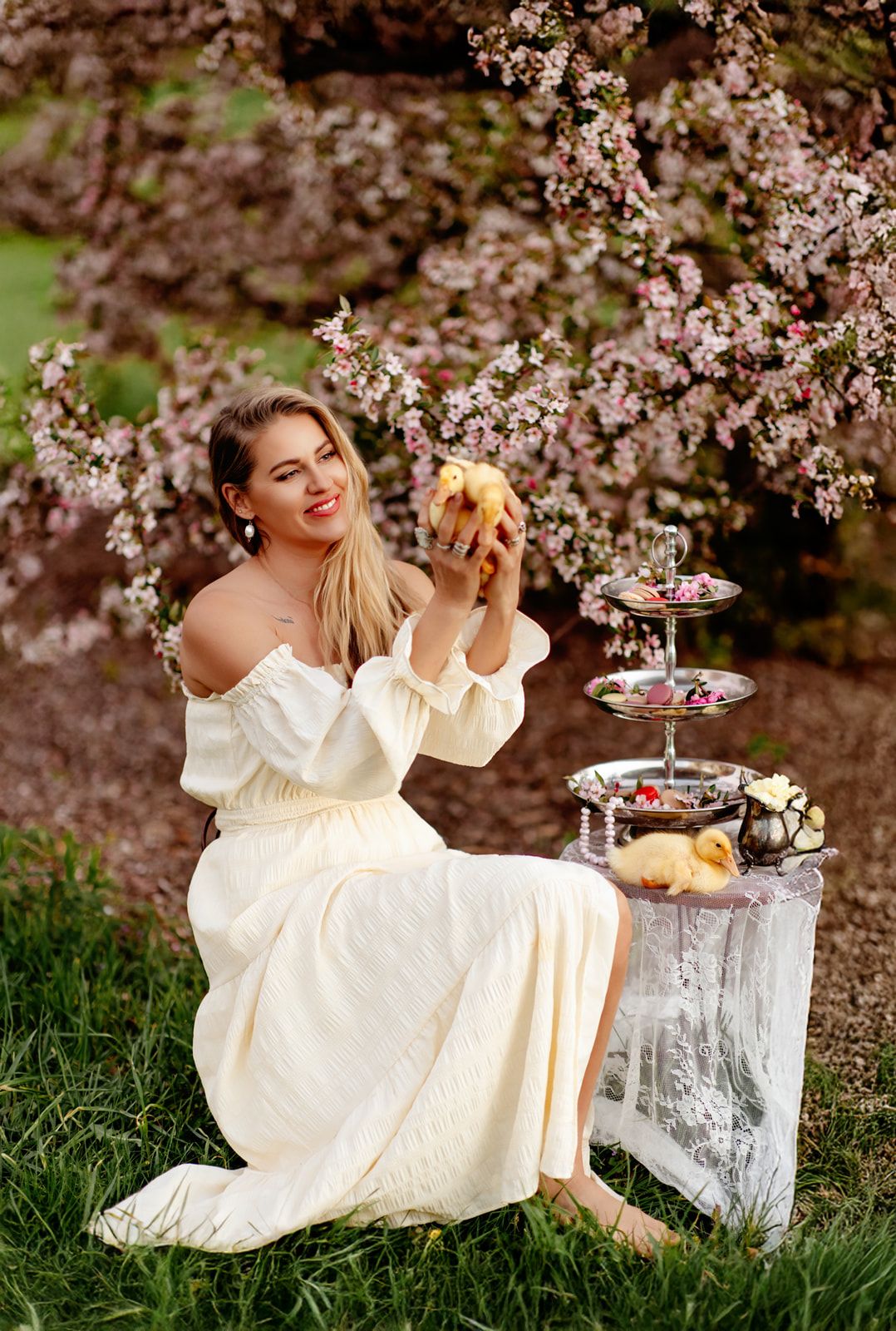 Woman in white off-shoulder dress holds chick near tiered dessert stand under pink blossoming tree.