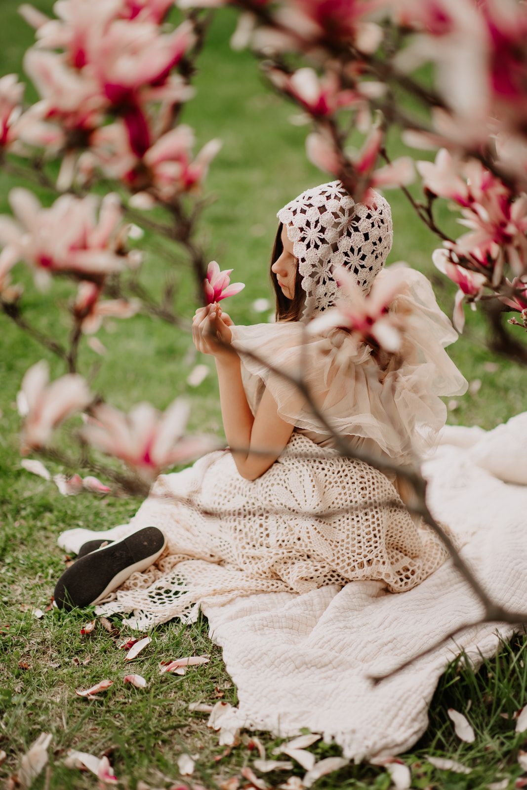 Girl in crochet bonnet and beige dress sits on blanket under blooming magnolia tree holding pink flower.