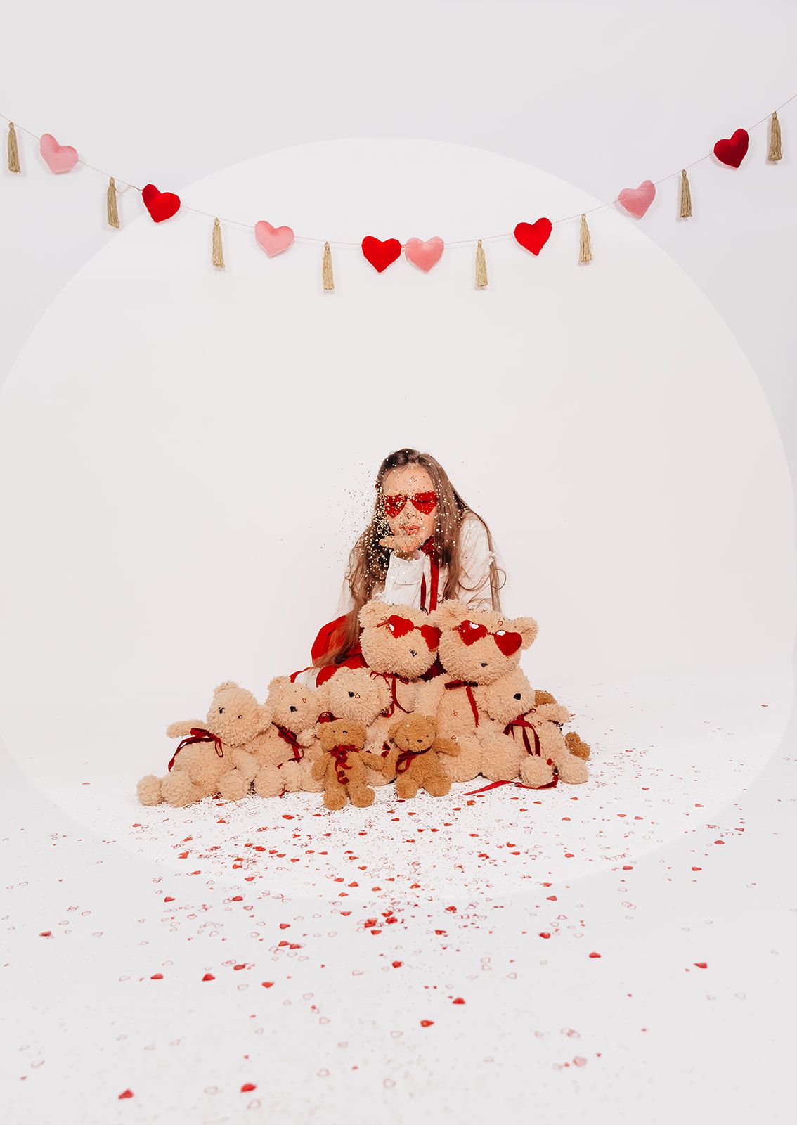 Girl surrounded by teddy bears with red bows, wearing heart sunglasses, under a heart garland with confetti on white background.