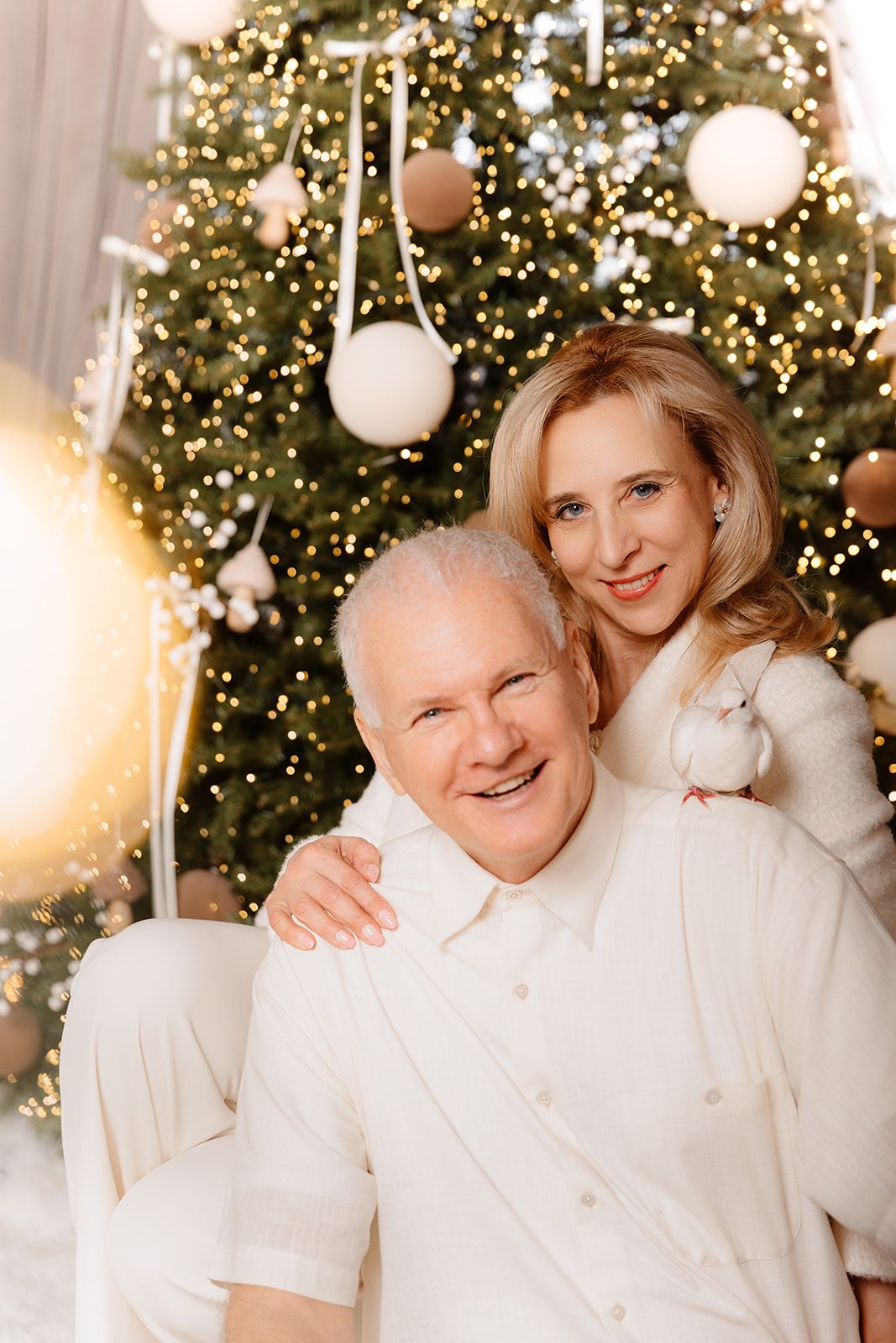 Smiling couple in white outfits embracing in front of a decorated Christmas tree with warm golden lights.