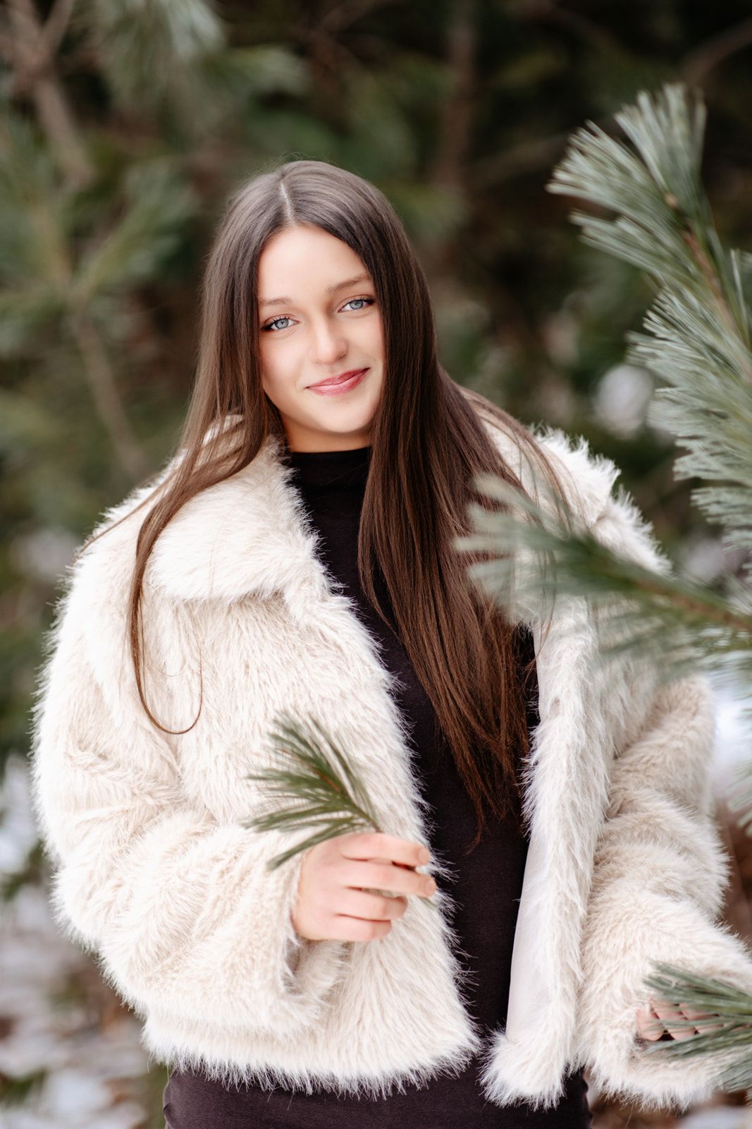 Young woman with long dark hair smiles in white faux fur coat, holding pine branch in snowy winter forest.