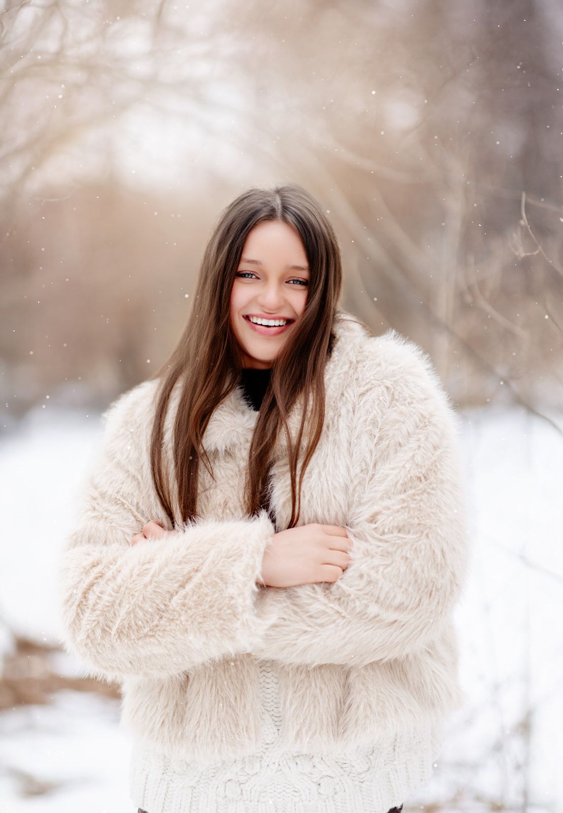 A smiling teenage girl with long brown hair wearing a cream faux fur coat stands joyfully in a snowy winter landscape.