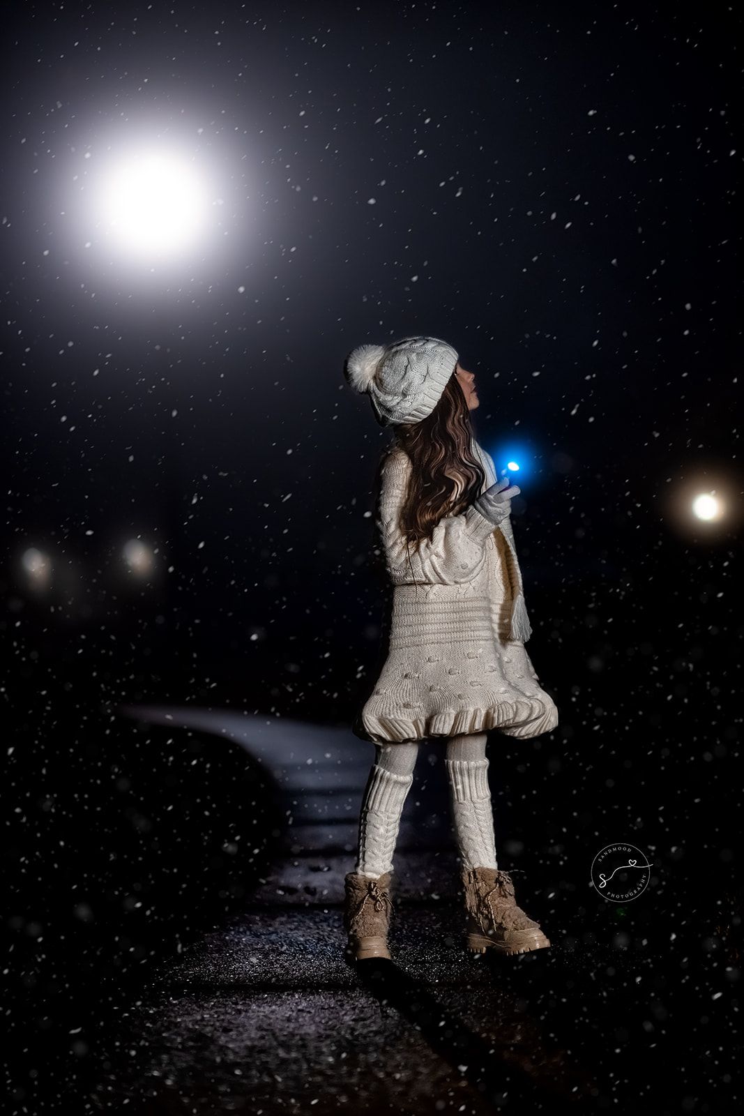 A young girl in winter clothes and knit hat stands on a dark path surrounded by falling snow and glowing lights at night.