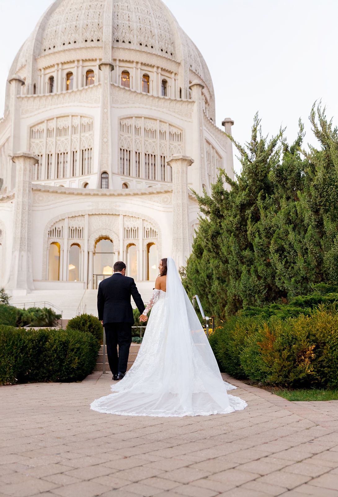 Bride and groom holding hands walking toward a grand white domed Bahai Temple surrounded by lush greenery.