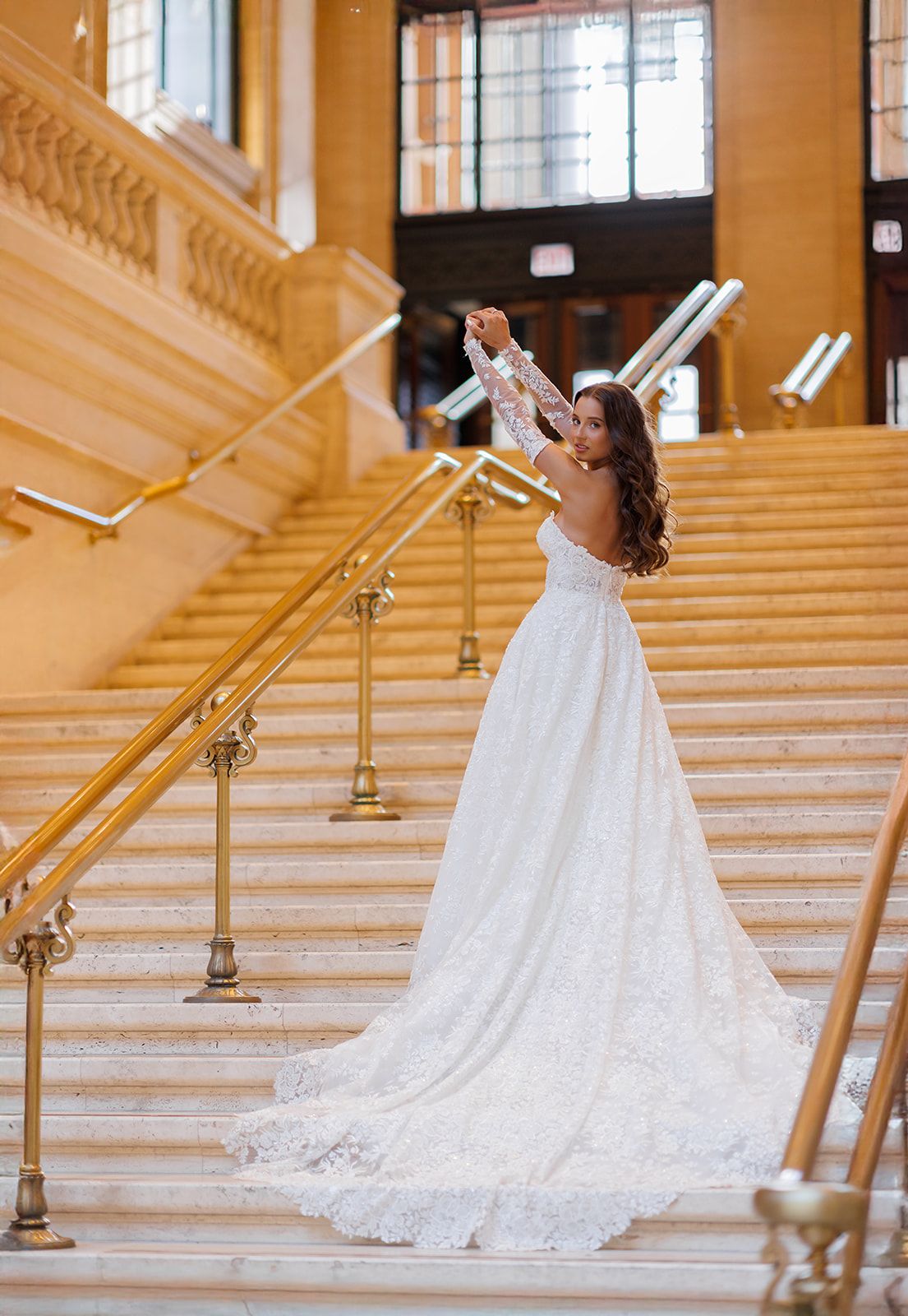 Bride in a white lace gown with a long train poses on grand staircase in an elegant venue.