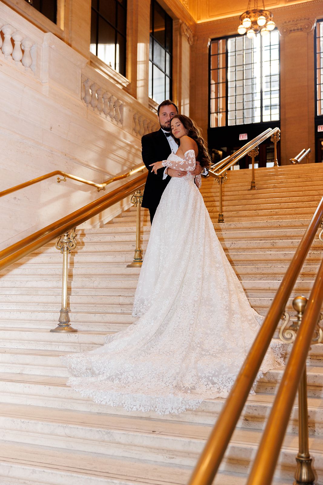 Bride and groom pose on grand marble staircase with gold railings, bride wearing elegant lace off-shoulder wedding gown.