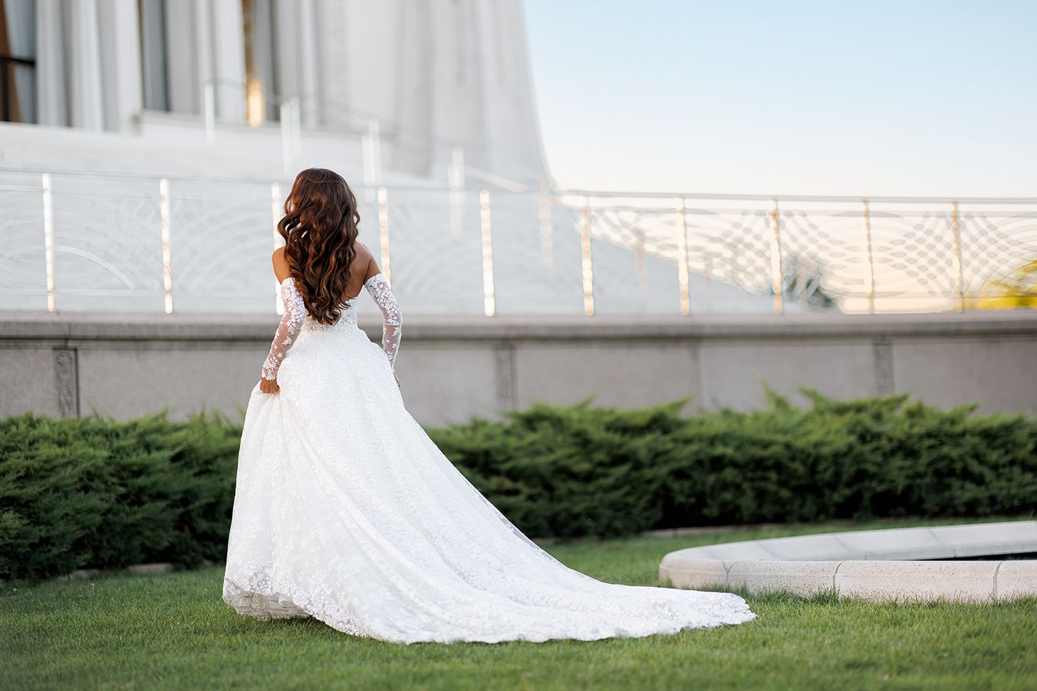 Bride in lace long-sleeve wedding gown with train stands before a grand white building at dusk.