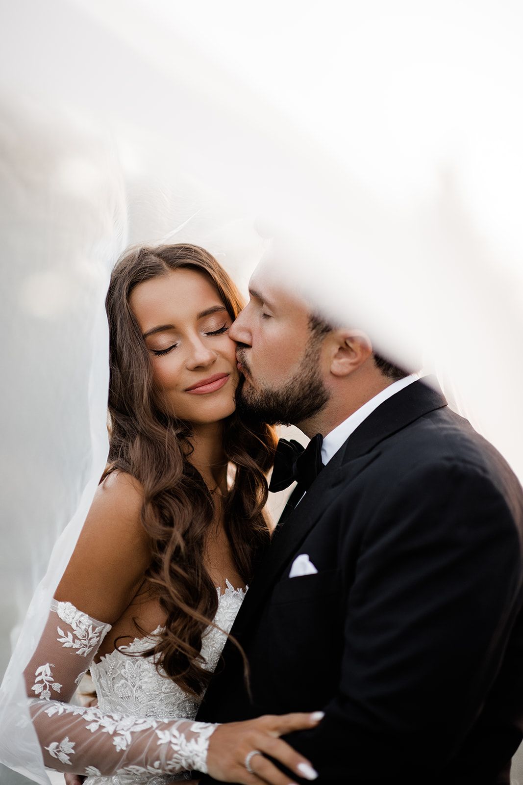 A groom in a black tuxedo gently kisses his bride on the cheek as she smiles, framed by a flowing white veil.