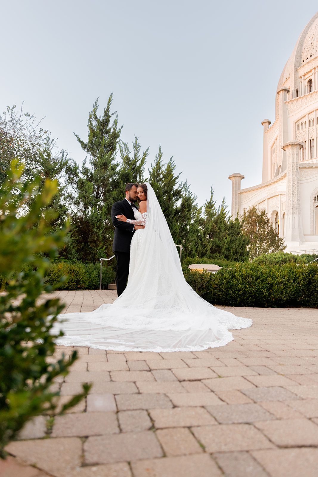 Bride and groom embrace in lush garden courtyard beside the elegant white Bahai Temple on their wedding day.