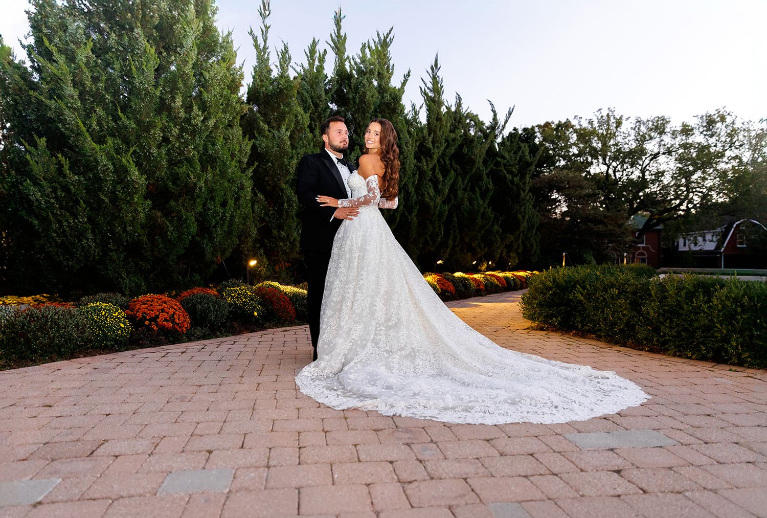 Bride and groom pose on brick patio surrounded by lush greenery and flowers at dusk in elegant wedding attire.