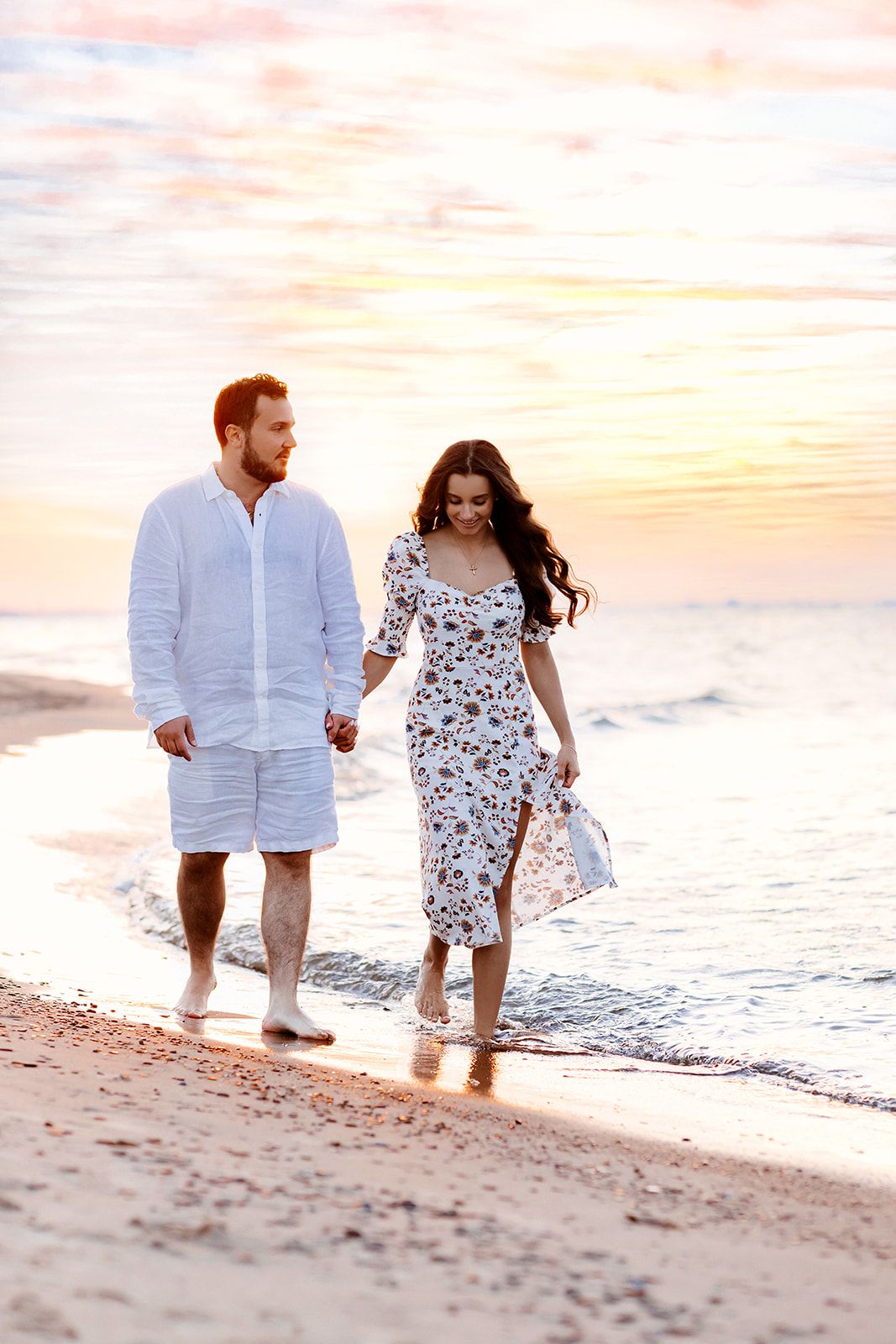 Romantic couple holding hands walking along shoreline at colorful sunset, woman in floral dress.