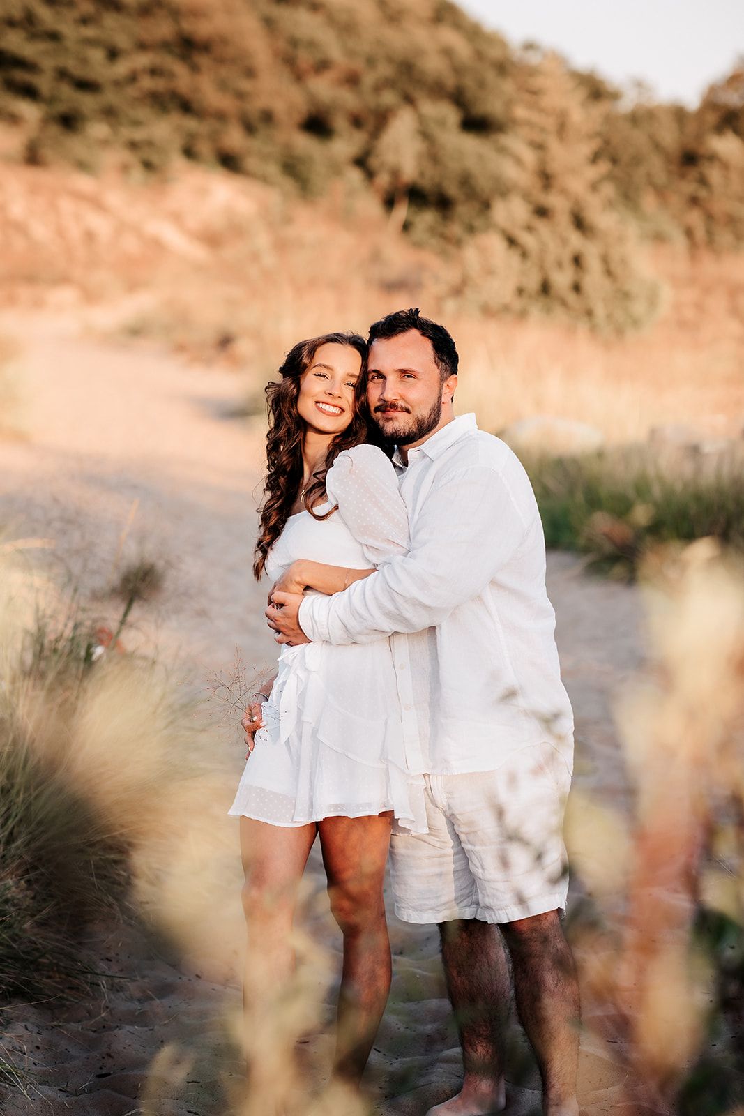 Romantic couple embracing on sandy beach path surrounded by golden dune grass at sunset.