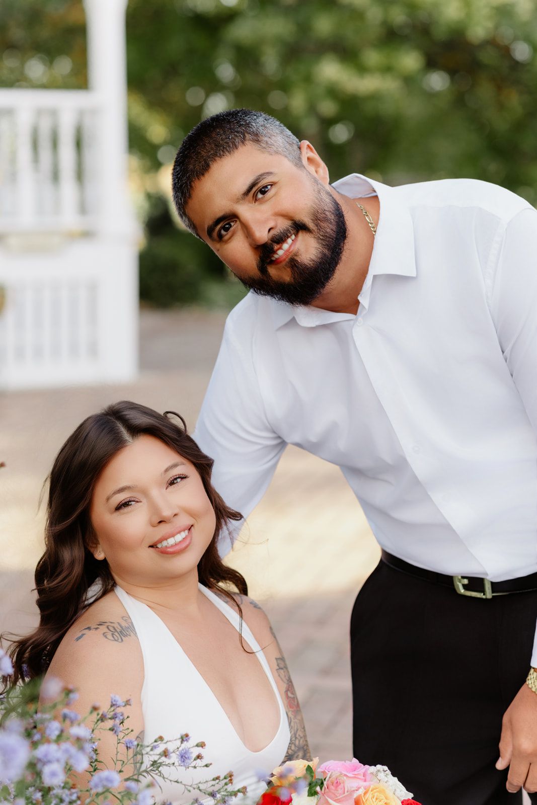 Smiling bride and bearded groom pose together near colorful floral arrangement at outdoor venue.