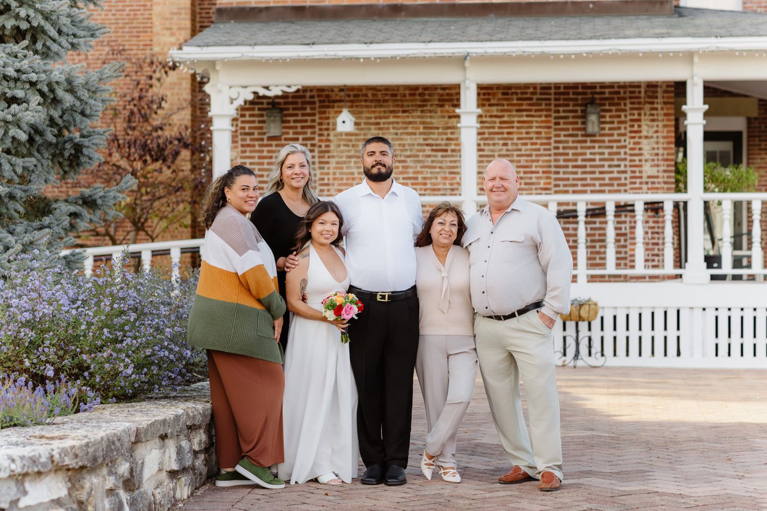 Wedding couple poses with family outside brick house with white porch, bride holds colorful bouquet.