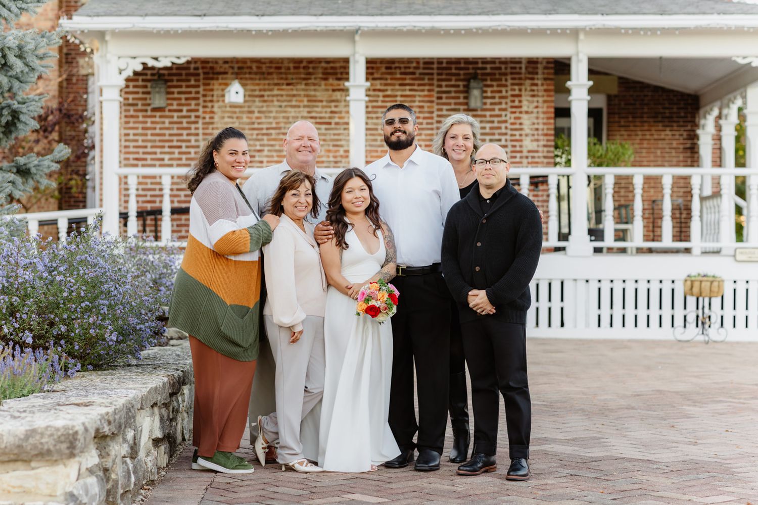 Wedding party of seven poses together in front of a brick house with white porch after the ceremony.