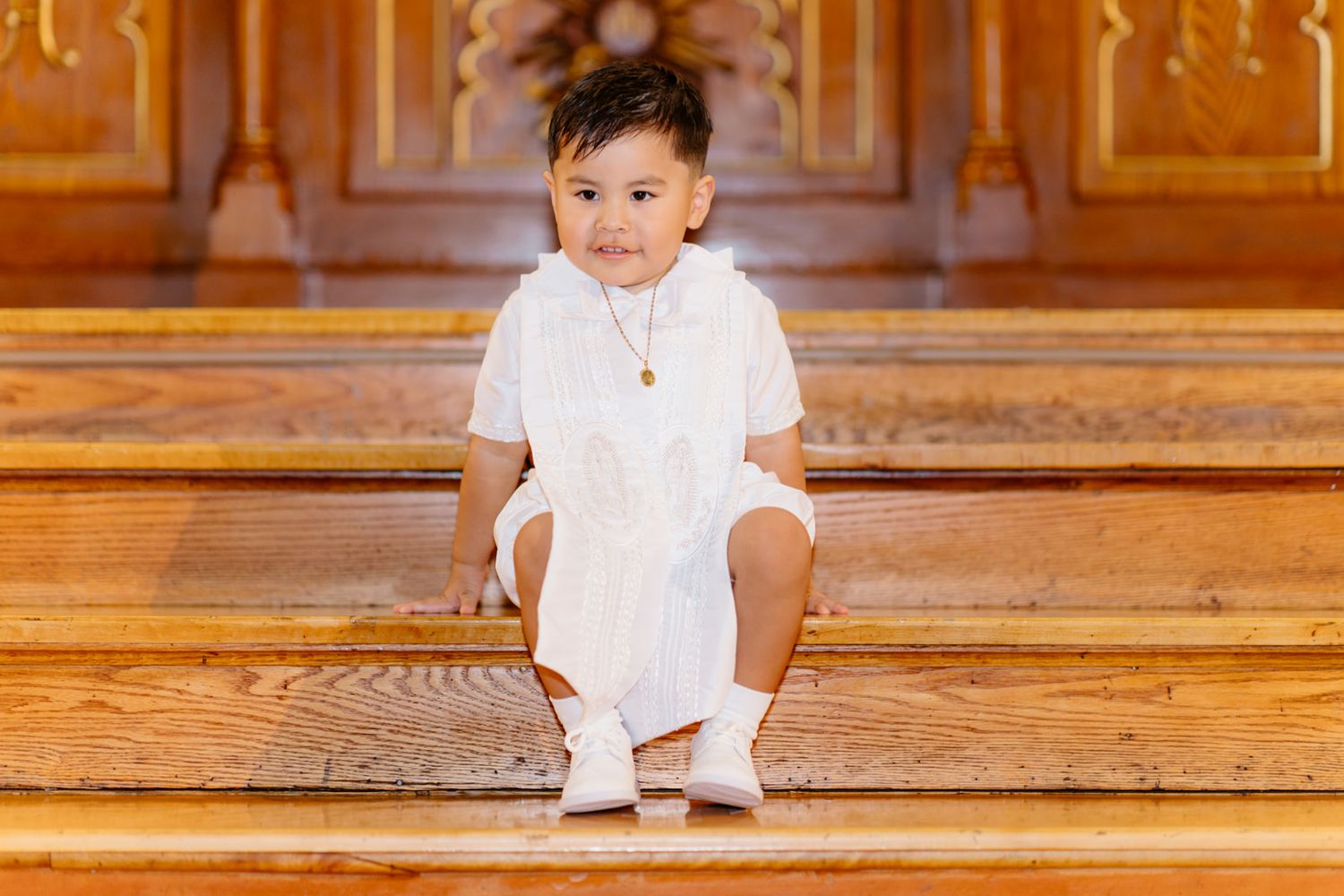 Young boy in white outfit sitting on wooden church steps, wearing a gold necklace, dressed for a special occasion.