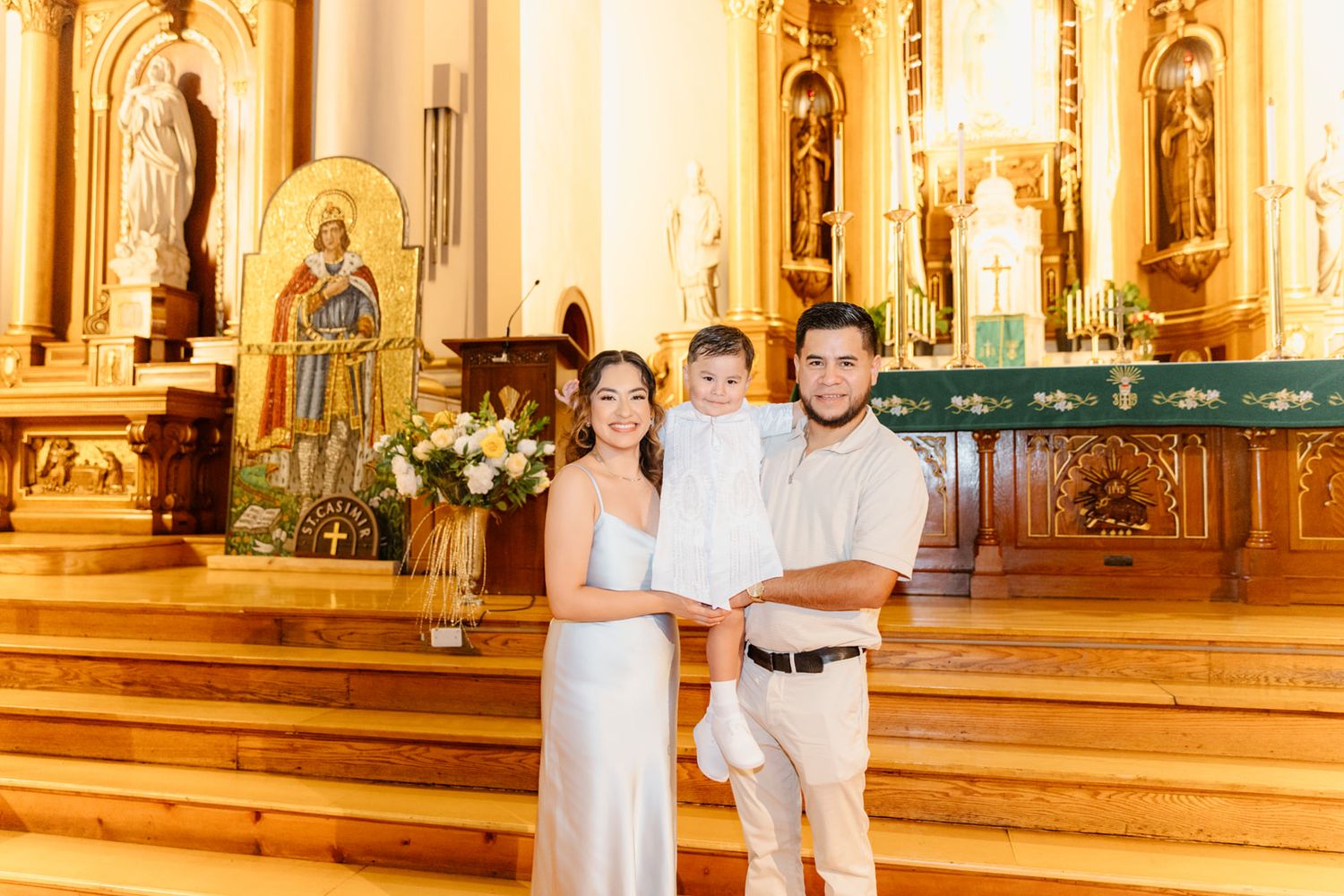 Family poses at church altar during baptism ceremony, parents holding baby in white outfit.