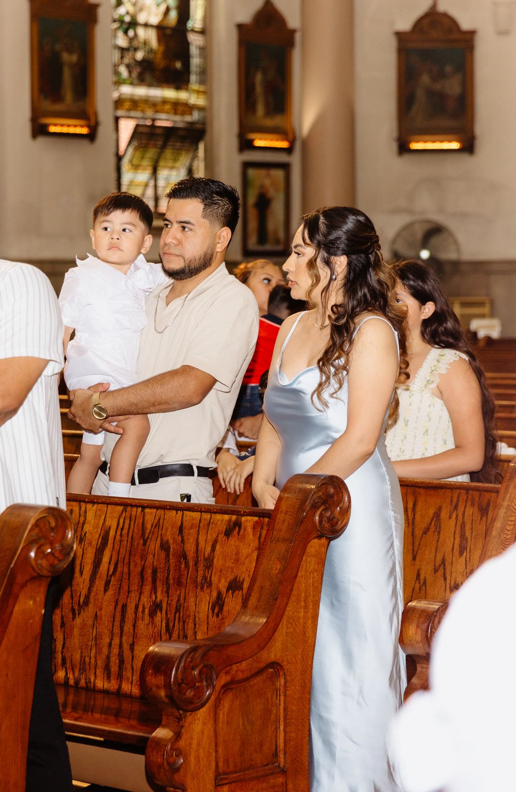 Family standing in church pews during a baptism ceremony, man holding toddler in white outfit, woman in blue satin dress.