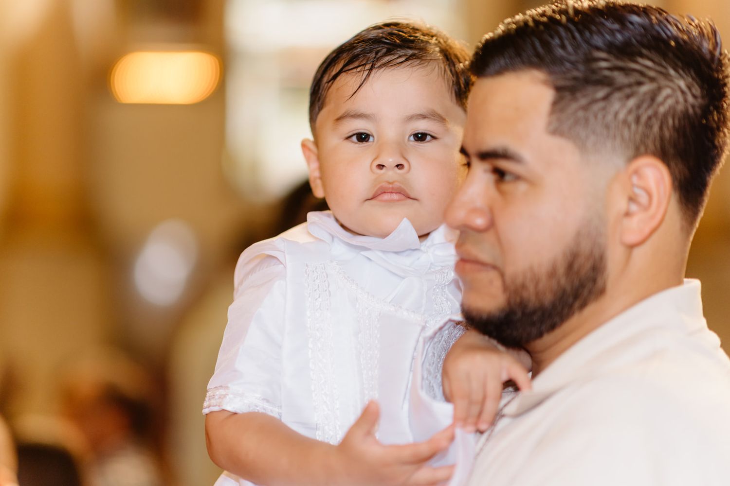 A man with a beard holds a young toddler dressed in white at what appears to be a baptism ceremony.