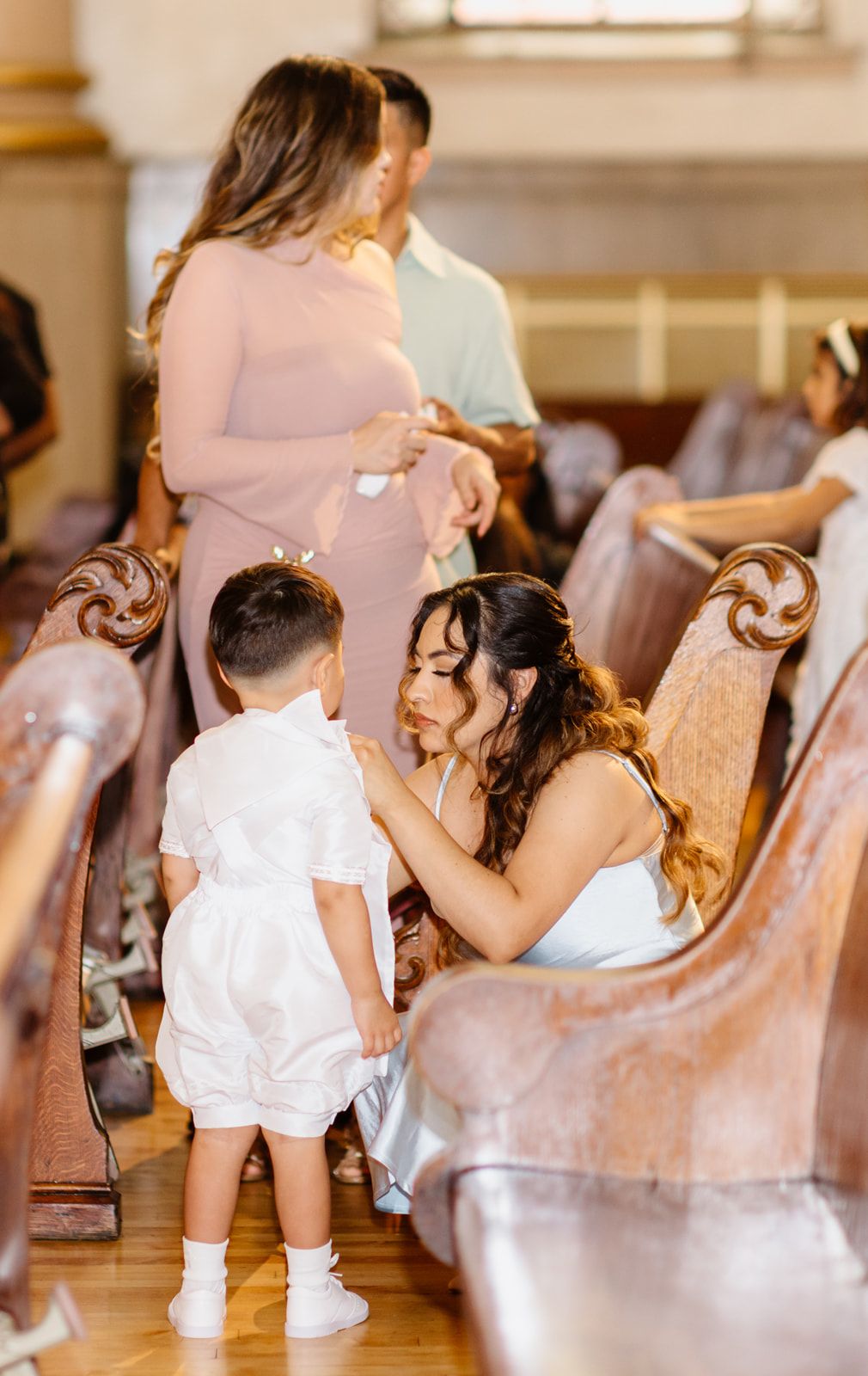 Woman in blue dress adjusting young boy's white outfit in church pew during a formal religious ceremony.