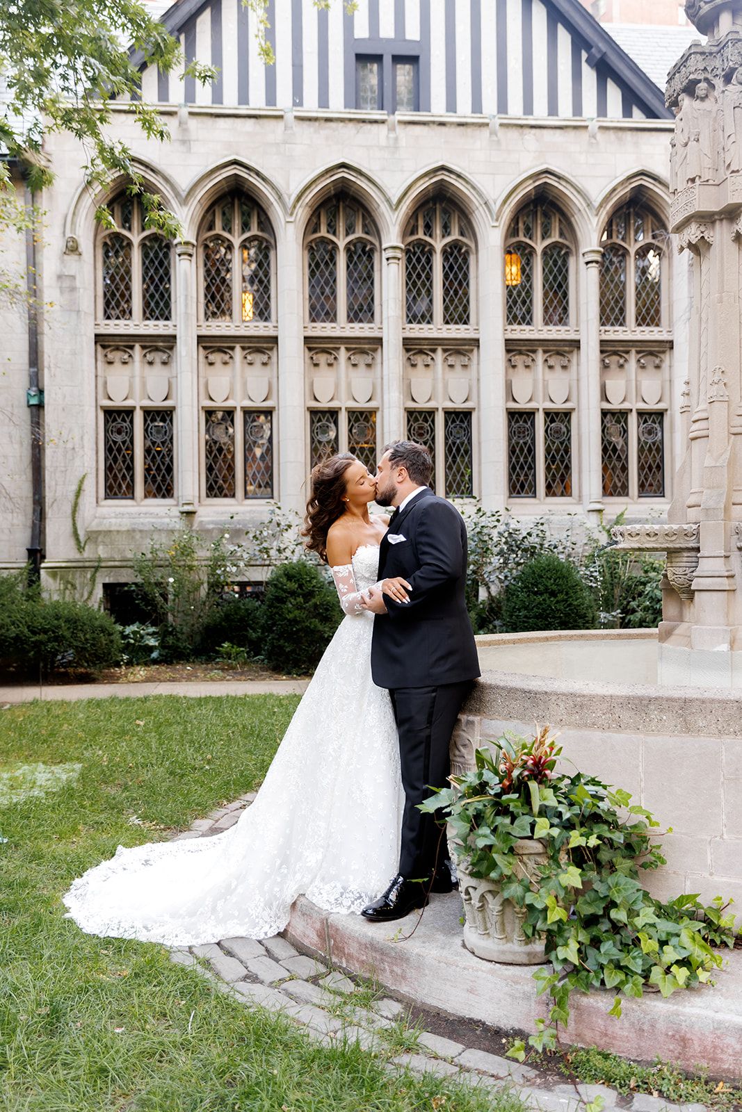Bride and groom share a romantic kiss outside a Gothic stone building with arched windows and lush ivy.