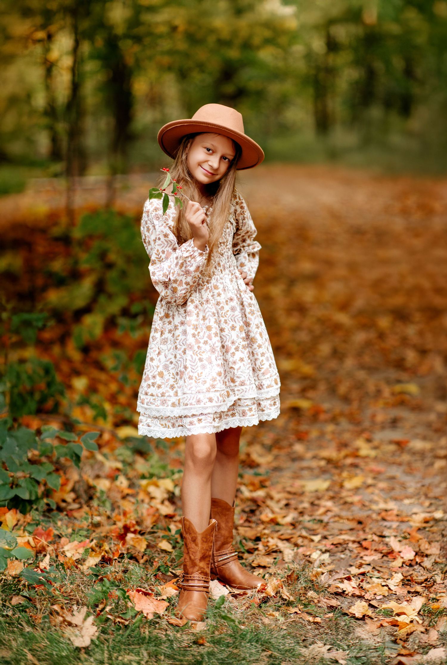 A young girl in a floral dress, tan hat, and brown cowboy boots poses on an autumn leaf-covered path.
