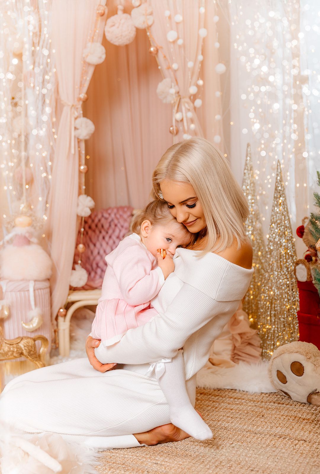 Blonde woman in white off-shoulder dress holds smiling baby in pink outfit amid festive holiday decor with fairy lights.