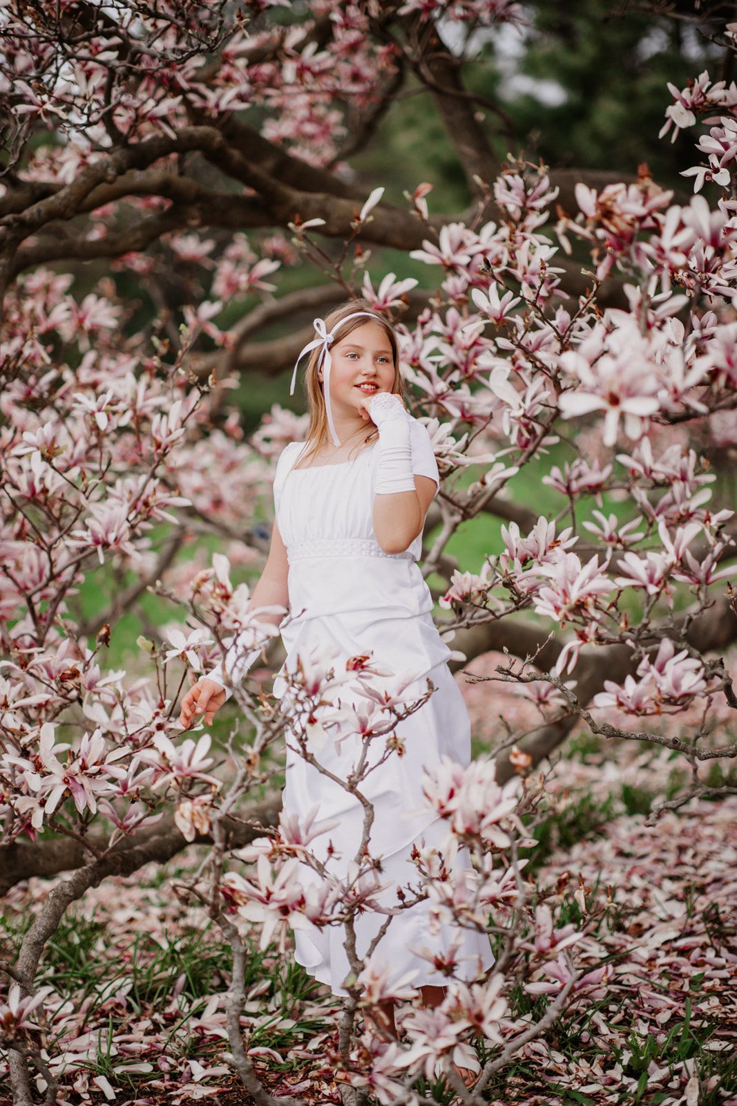 Smiling woman in white dress posing among pink magnolia blossoms in a flowering spring garden.