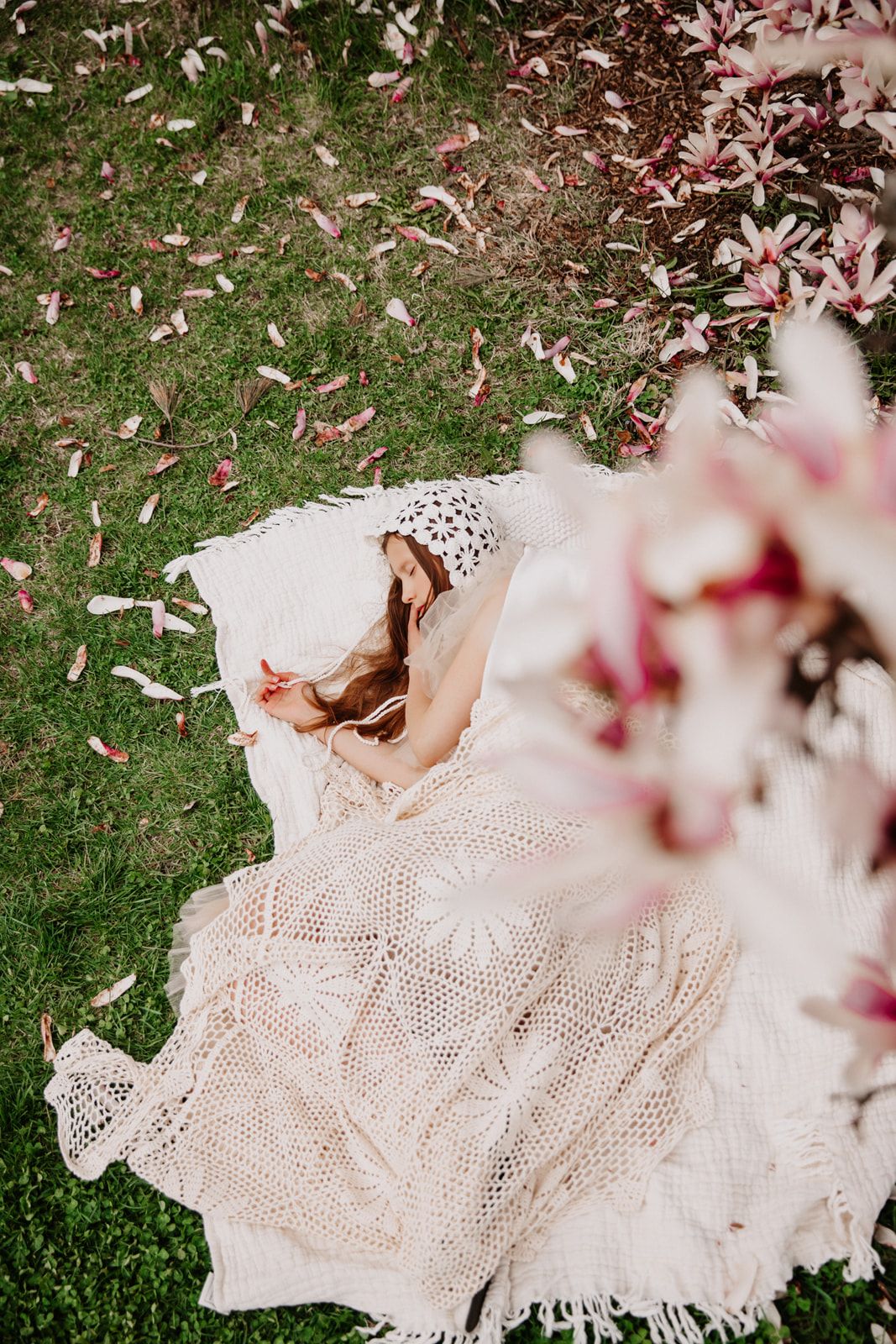 Aerial view of a woman lying on a crochet blanket on grass surrounded by pink magnolia petals.