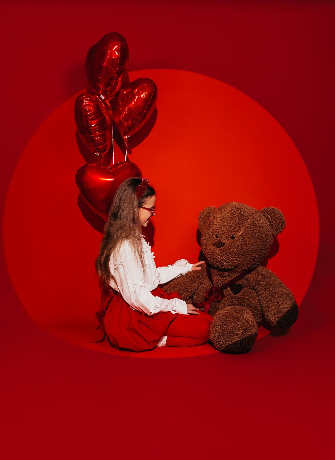 Young girl in red skirt sitting with large teddy bear and red heart balloons on vibrant red background.