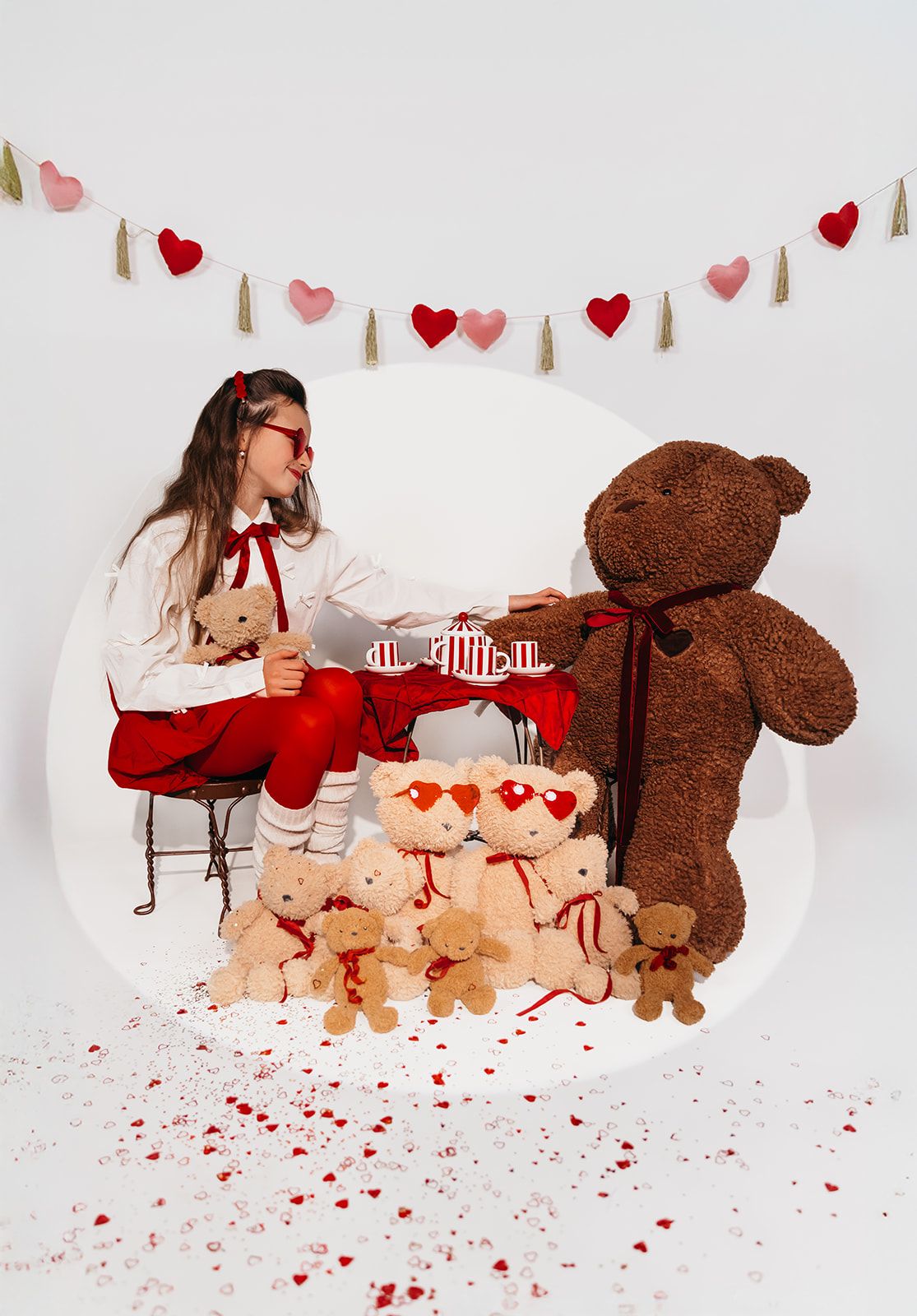 Girl in red glasses sits at tea table with large brown teddy bear and small Valentine bears, heart garland backdrop.