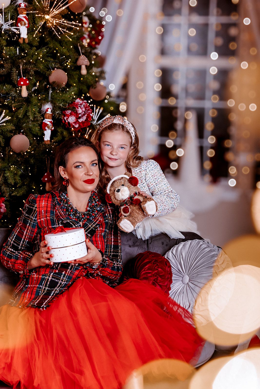 Mother and daughter in matching red plaid outfits pose with gifts by a decorated Christmas tree.