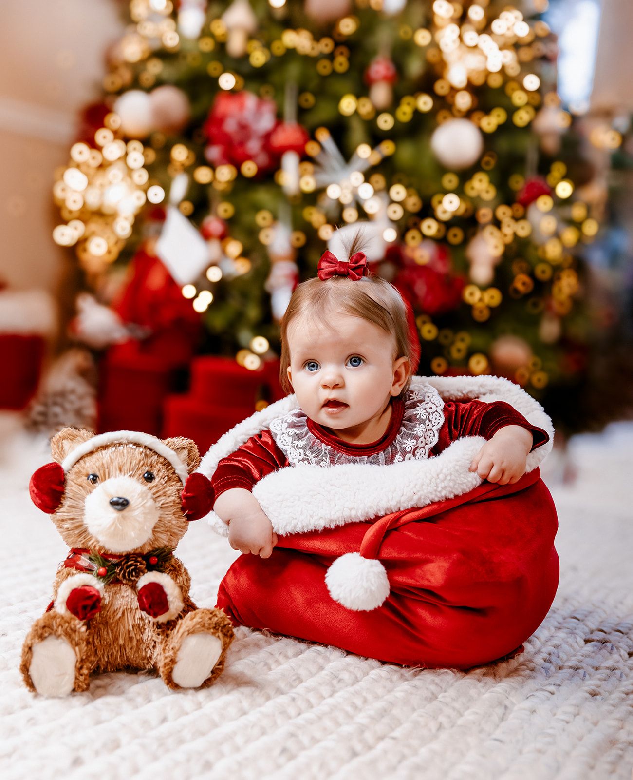 Baby in red velvet outfit sits in Santa sack beside teddy bear in front of decorated Christmas tree.
