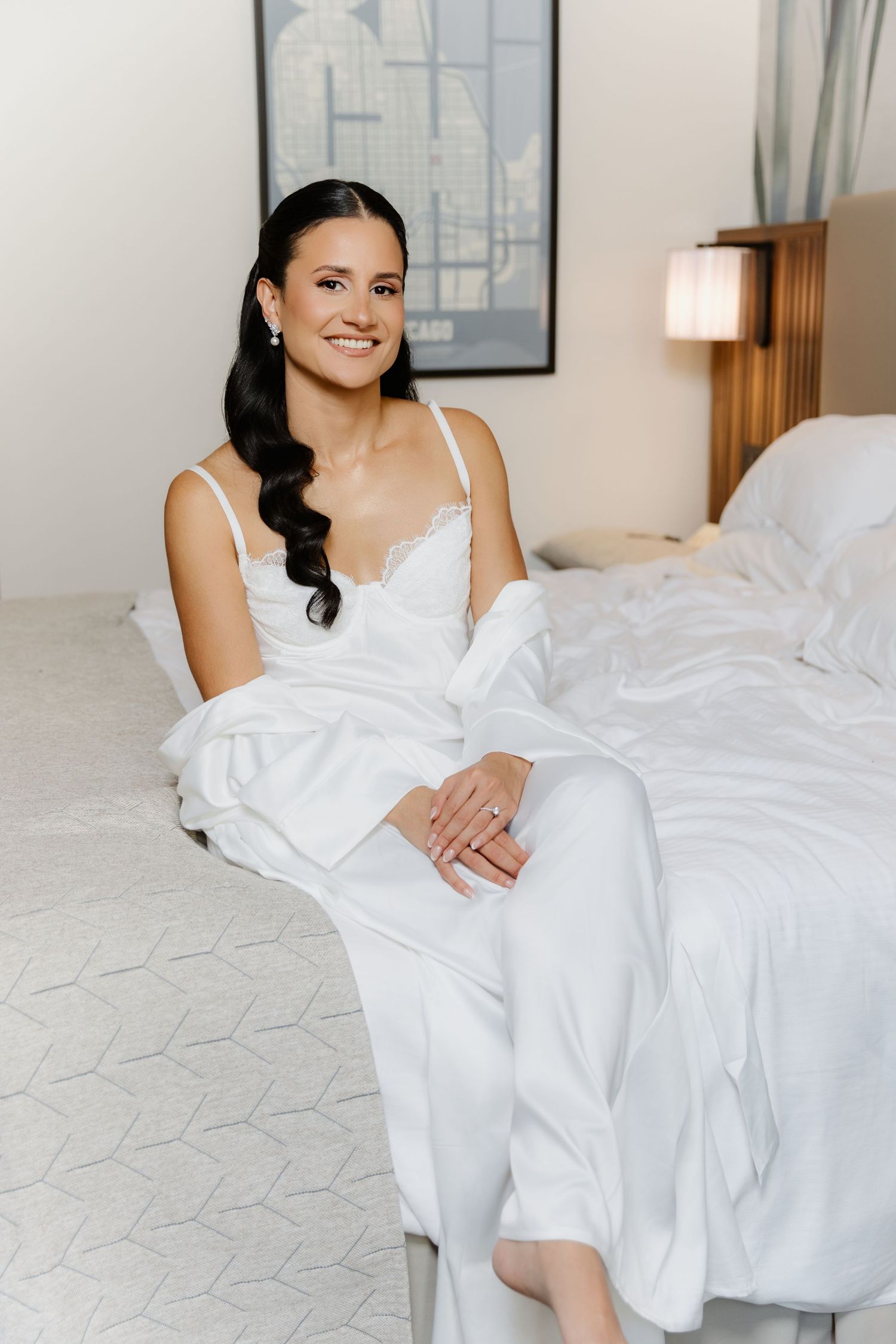 Elegant bride with dark wavy hair smiles in white lace slip dress and satin robe, seated on white hotel bed.