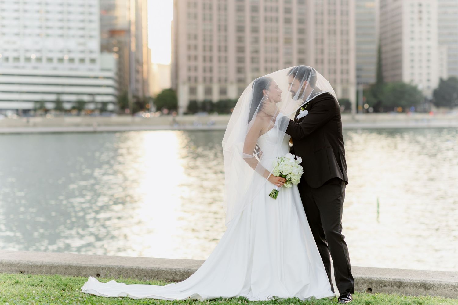 Bride and groom sharing romantic kiss under veil by waterfront with city skyline backdrop.