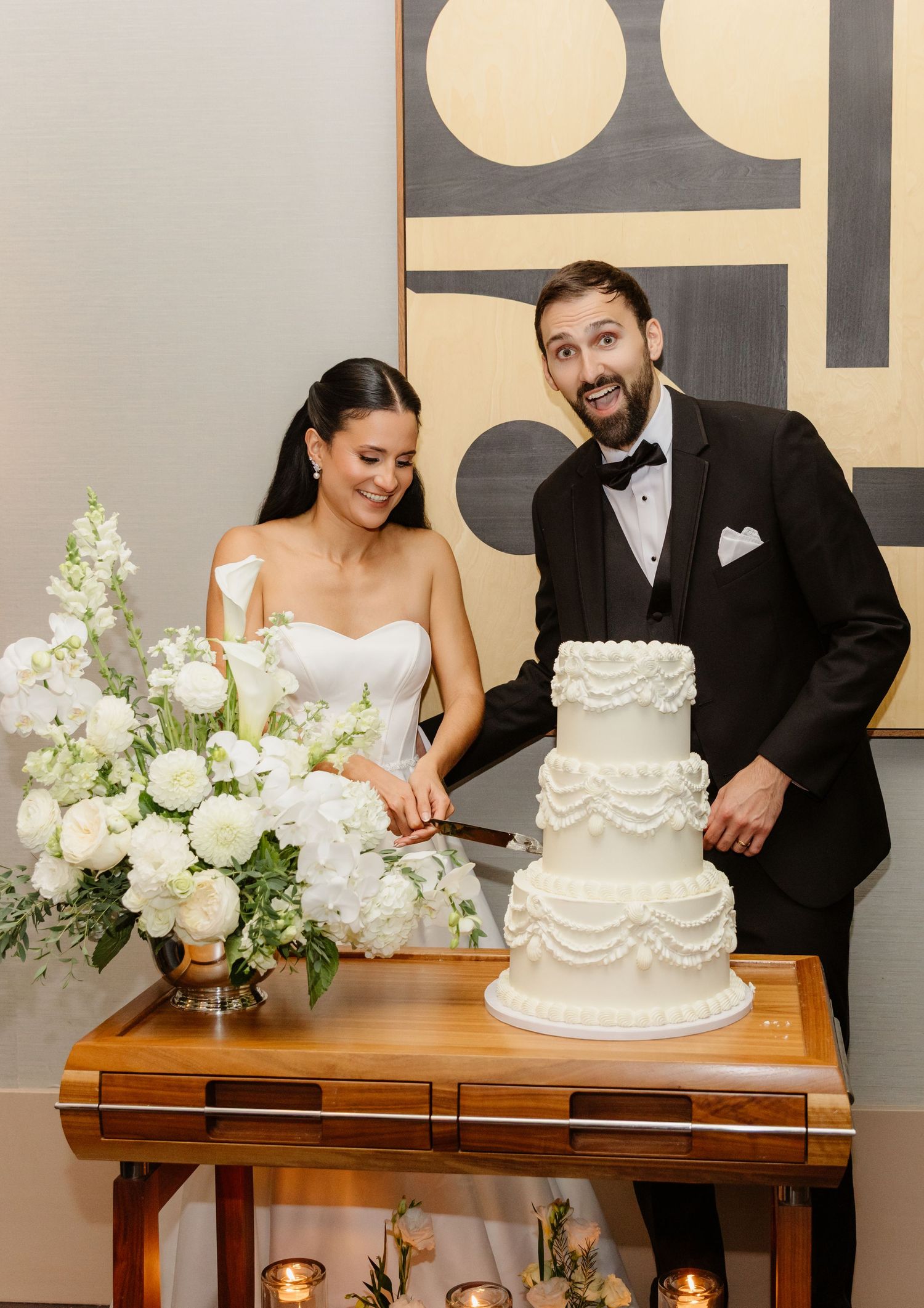 Excited groom and smiling bride cutting their ornate white ruffled tiered wedding cake at their reception.
