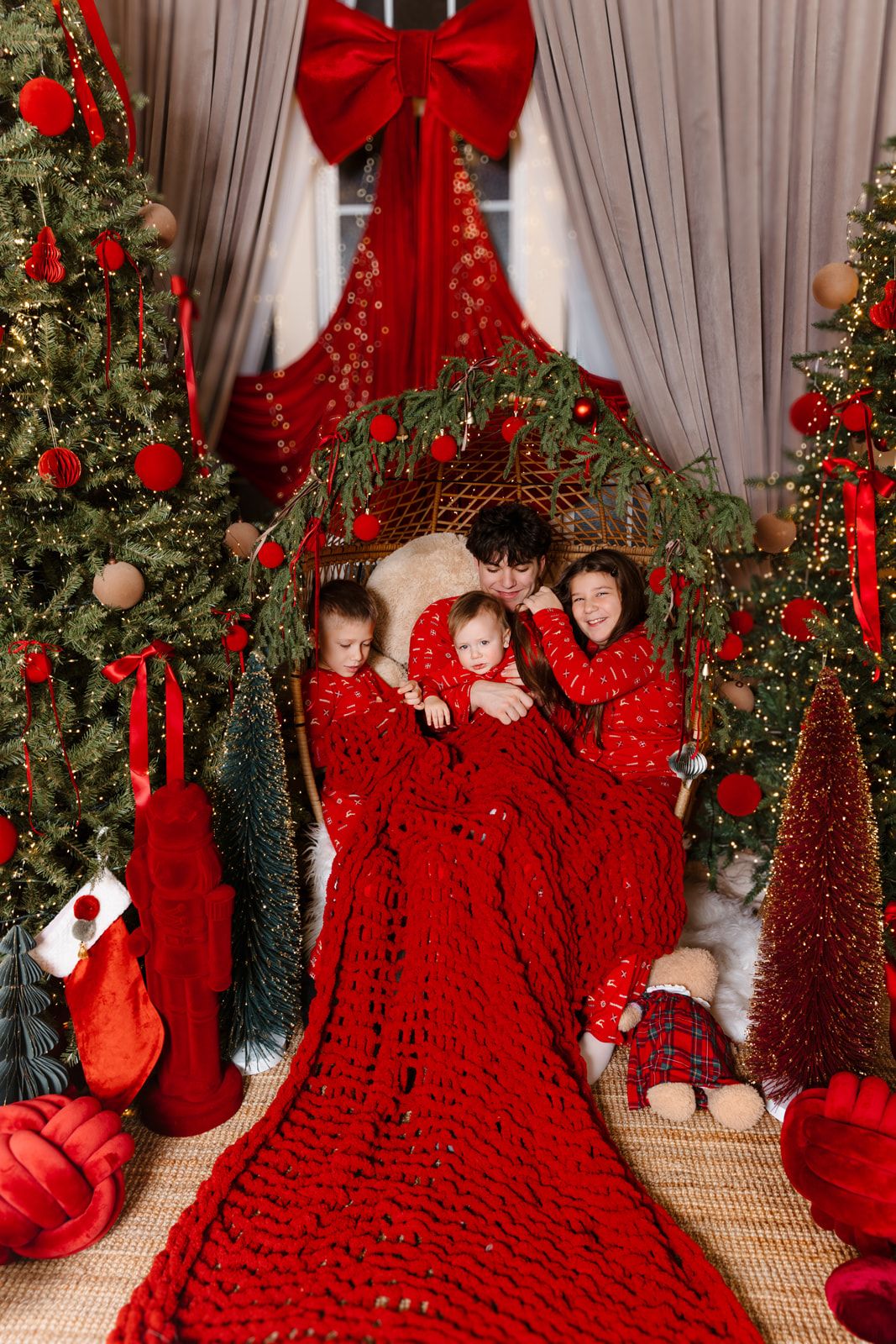 Family in red pajamas cuddles under a chunky knit blanket surrounded by Christmas trees with red ornaments and holiday decor.