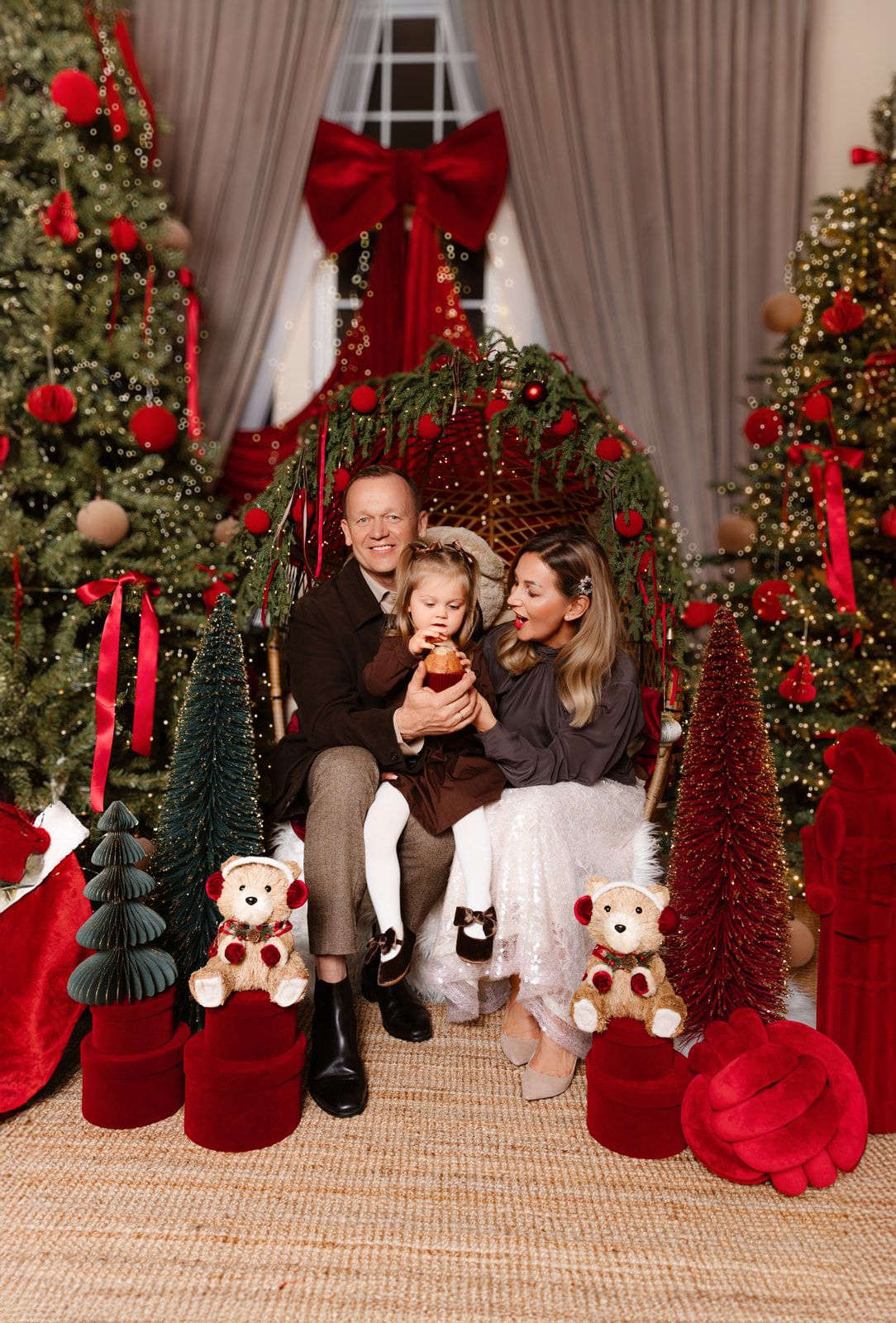Family of three poses with Christmas trees, red bows, teddy bears, and holiday decor in a festive portrait.