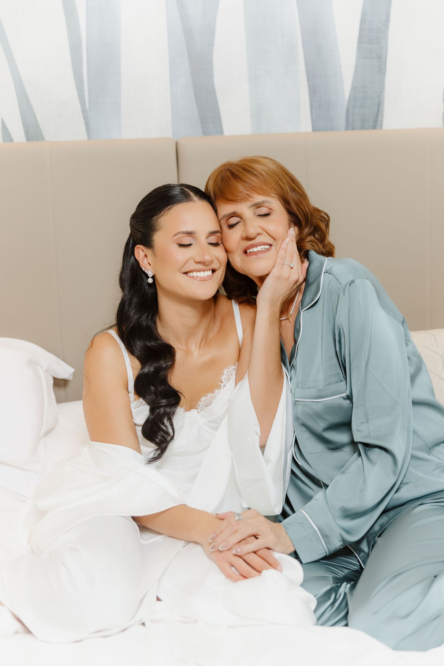 A bride in white lingerie and her mother in teal pajamas sit together on a bed, smiling warmly on a wedding morning.