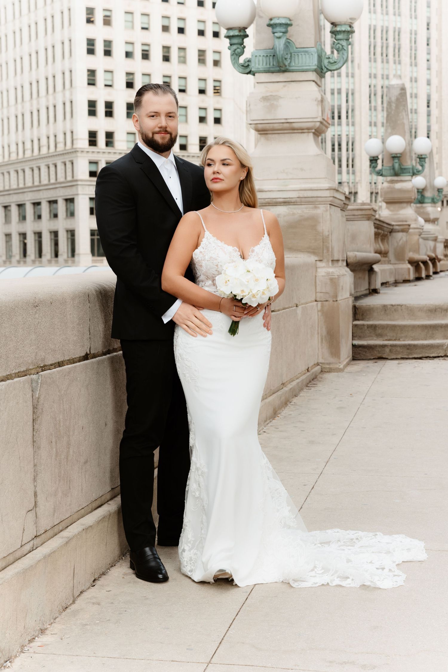 Elegant bride in lace gown holds white bouquet as groom in black tuxedo poses on Chicago bridge.