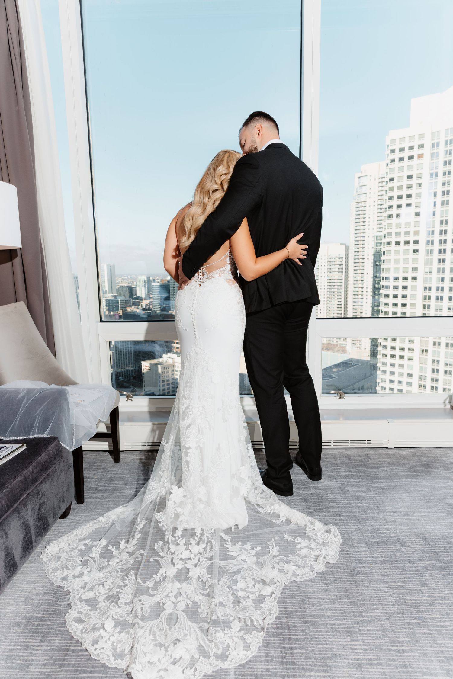 Rear view of bride in lace mermaid gown with floral train and groom in black suit embracing before city skyline windows.
