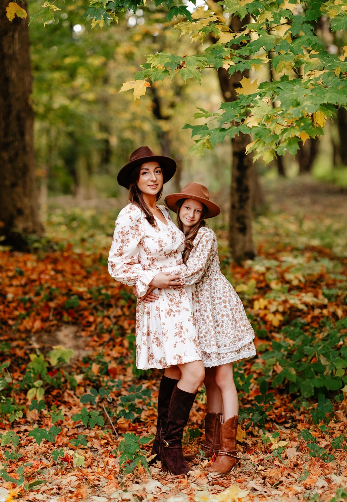 Mother and daughter in matching floral dresses and wide-brim hats embracing in colorful autumn forest.